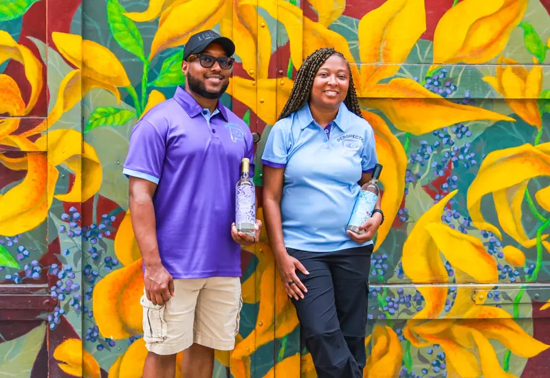 Crystal and Roger, founders of Perspection Spirits, holding bottles in front of a floral mural